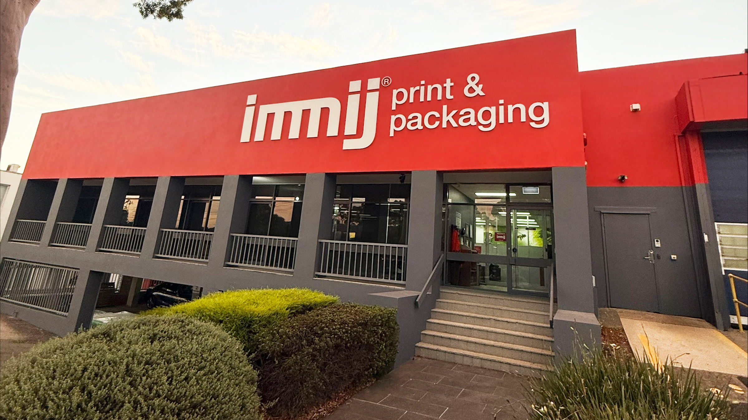 A building with a red and grey exterior displays the sign immij print & packaging above its entrance, showcasing print and packaging Australia expertise, surrounded by greenery and a paved walkway.